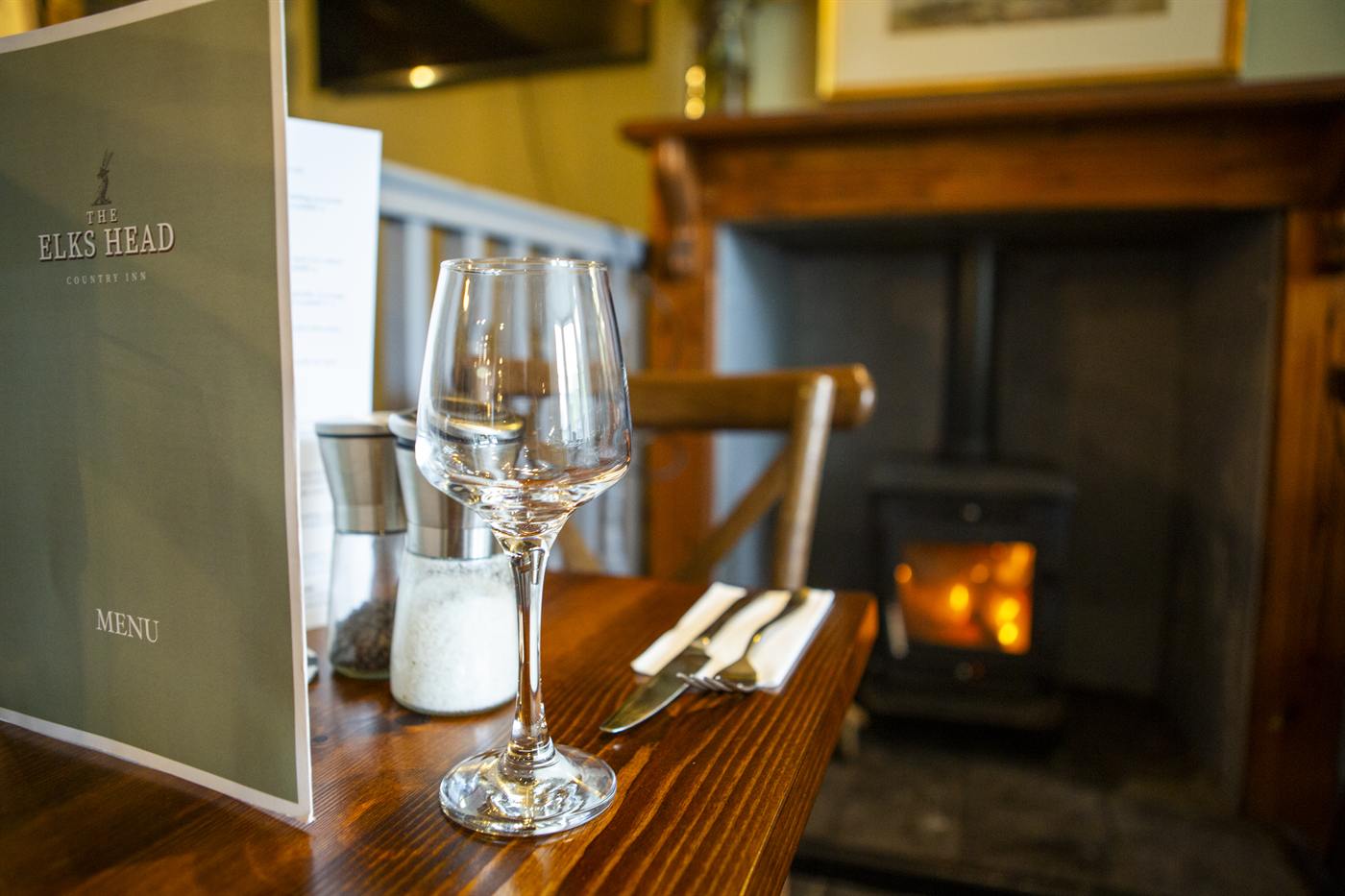 A table set with a wine glass and menu beside the log burner