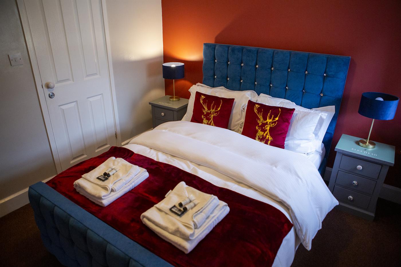 A double bedroom with a blue velvet headboard and red Elks Head cushions